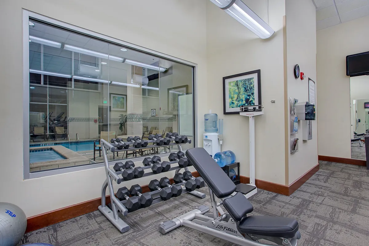 A motivating fitness center with a dumbbell rack, weight bench, water cooler, and a window view of the pool, blending wellness and social opportunity.