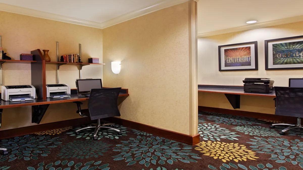 A productive business center featuring desks with computers, a colorful carpet, and inspirational wall art. The scene captures a welcoming environment designed for work, study, or casual browsing, reinforcing the property's value for business and extended-stay travelers.