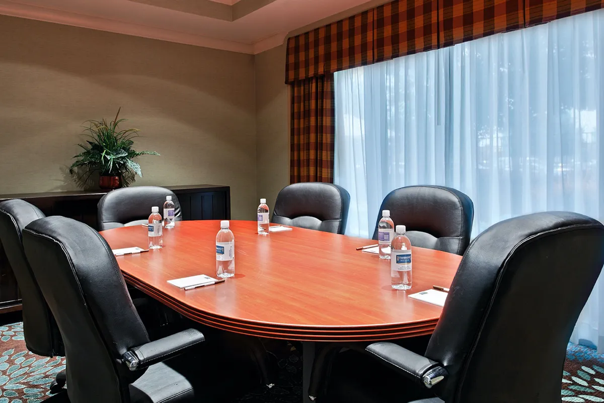 This image depicts a spacious interior with a large oval table surrounded by black leather chairs, complemented by water bottles and notepads, suggesting a communal meeting or gathering space. The atmosphere is focused and collaborative, with elements that hint at both productivity and the ability to host group activities.