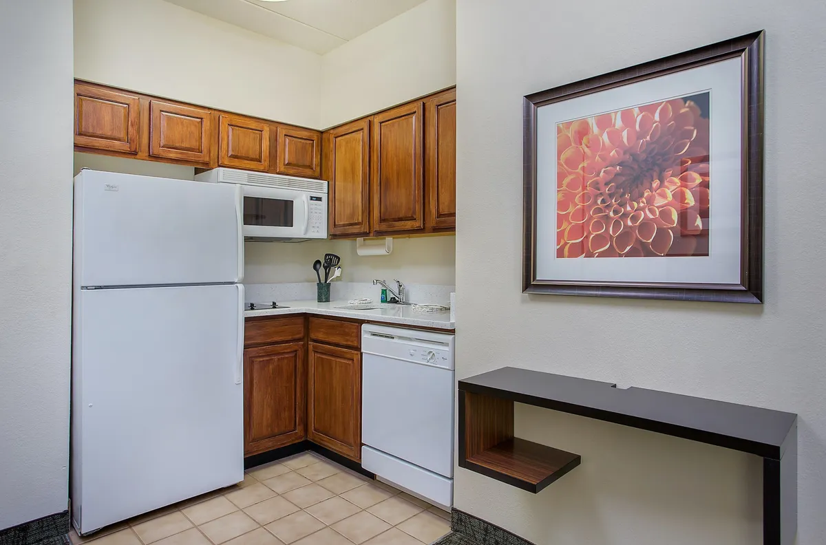 The image showcases a cozy studio kitchen area with warm wooden cabinets, gleaming white appliances, and elegant flower artwork adorning the wall. The inviting space is perfect for preparing meals, gathering with family, and enjoying comforting moments in Chattanooga's downtown core.