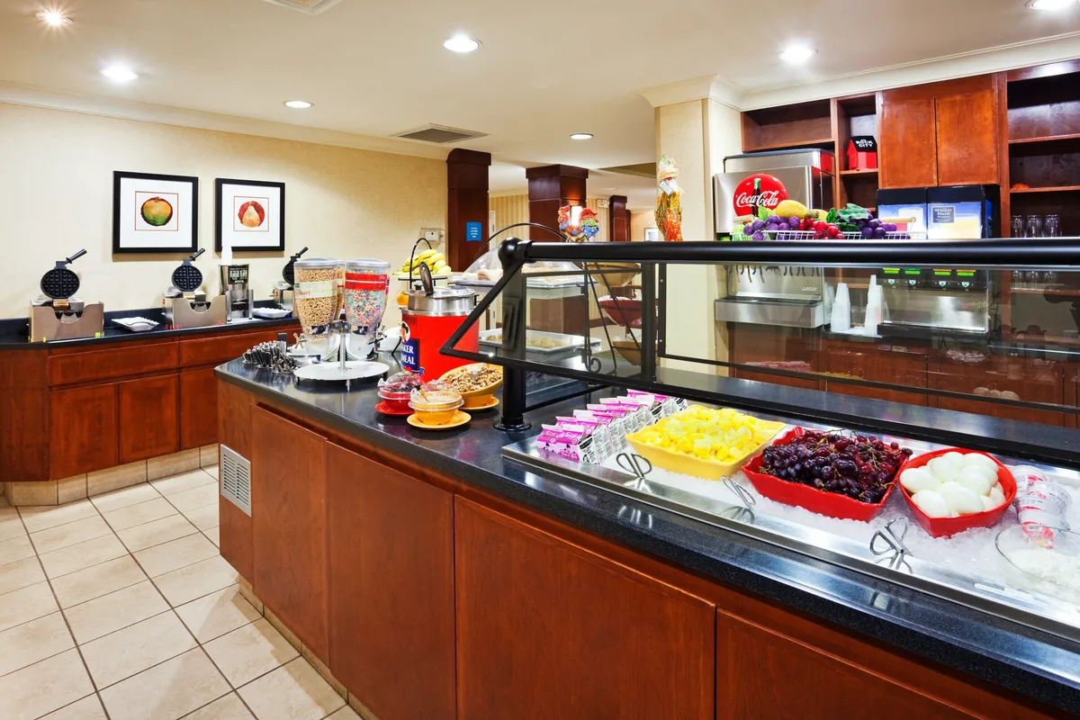 This image captures a breakfast bar stocked with a variety of breakfast offerings, including a fruit display, waffle maker, and a beverage station. Guests are seen casually helping themselves, setting a lively and welcoming morning mood.