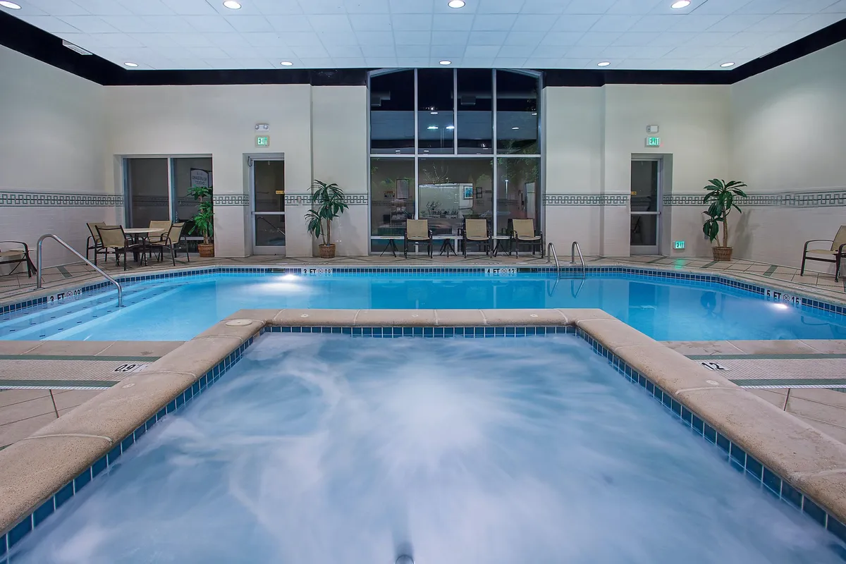 This image features a tranquil indoor pool area with a sparkling pool, inviting hot tub, lounge chairs, and lush plants. Soft lighting creates a welcoming ambiance ideal for relaxation or socializing.