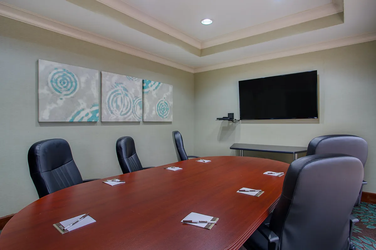 A modern meeting room with a large wooden conference table, comfortable black leather chairs, wall-mounted TV, and contemporary art, set up for business meetings and team discussions.