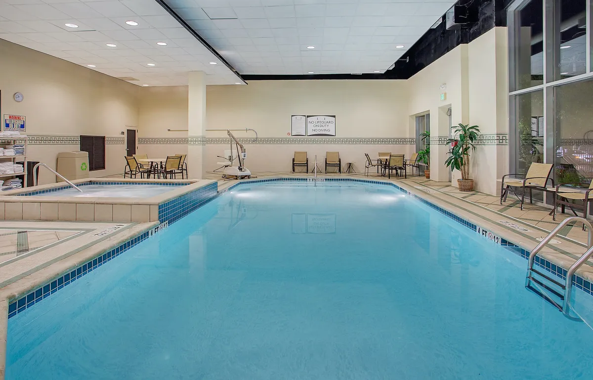 An interior shot of a tranquil indoor pool area, featuring lounge chairs alongside the pool, accented with potted plants and subtle signage. The space is designed for relaxation, socializing, or simply enjoying restful moments after a day of exploration.