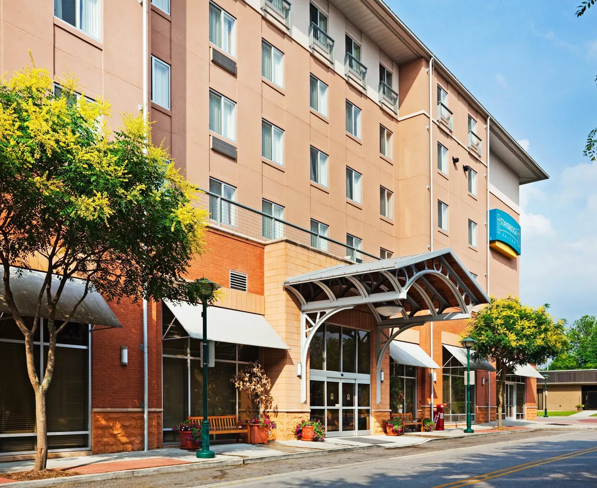 This shot features the hotel entrance with a stylish awning, large windows reflecting city life, colorful planters, and nearby seating areas. The vibrant, modern architecture and patio invite guests to relax outdoors, suggesting the ease of stepping out into the city or enjoying a coffee while soaking up the local atmosphere.