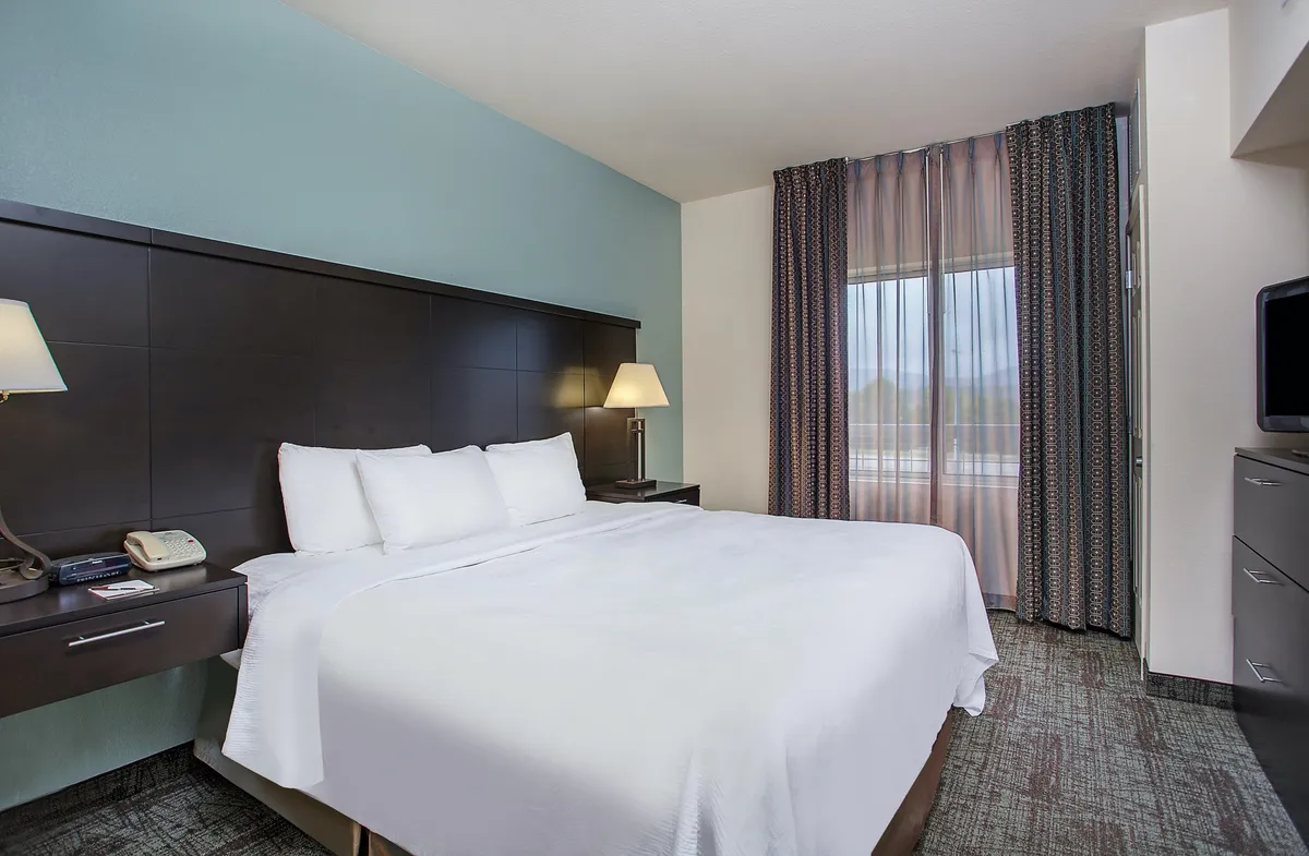 A bright, welcoming studio room with a neatly made Queen bed in crisp white linens, a nightstand with a lamp for evening reading, and a large window with soft curtains that let in natural light. The uncluttered space evokes a calm, restful atmosphere ideal for both relaxation and productivity.