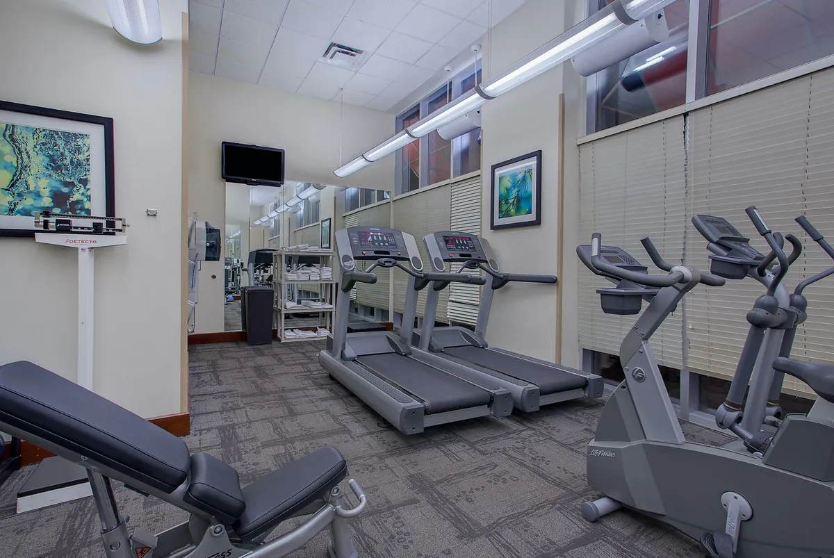 A modern fitness center equipped with treadmills, an elliptical machine, a weight bench, and large mirrors. The space is bright, clean, and inviting, with tasteful artwork on the walls adding character. The visible equipment suggests opportunities for both solo workouts and group wellness activities, emphasizing a communal and motivational atmosphere.
